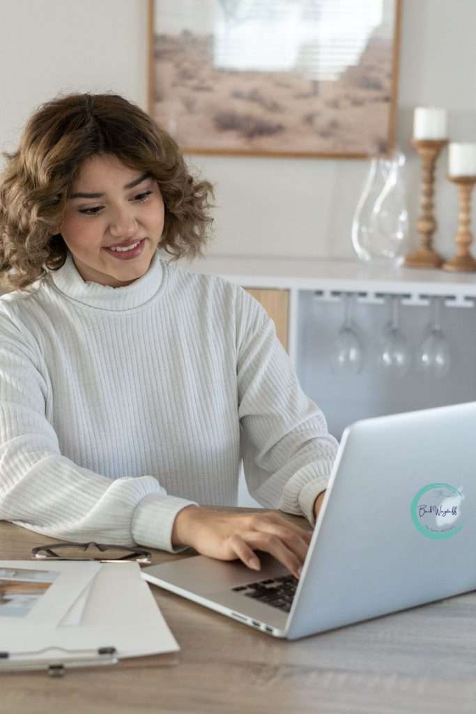 Asian woman in white sweater in home office typing at her laptop