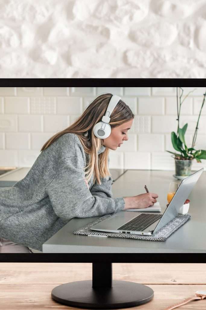 white background. Wooden desktop with black monitor. On the monitor is a photo of a woman in a grey sweater wearing white headphones and taking notes as she watches an onpage SEO workshop on the laptop in front of her.