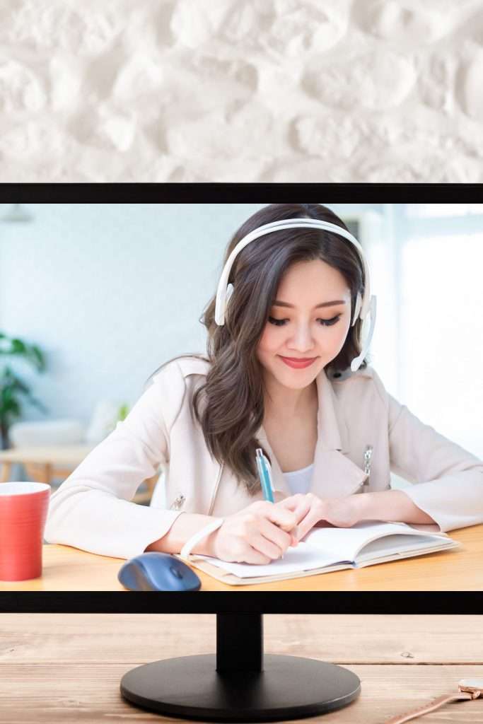 computer monitor sitting on a brown desk top with an image of a woman with dark hair sitting at a desk wearing headphones and taking notes from her online keyword research class