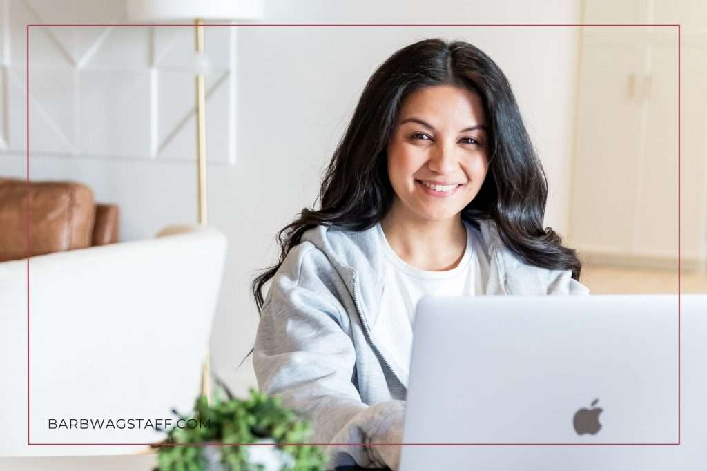 dark haired woman in grey hoodie smiling over top of her Mac computer. Green plant in the foreground. barbwagstaff.com written in the bottom left corner. 