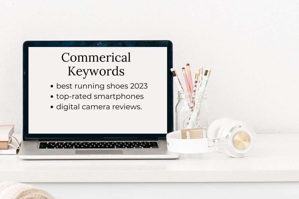 White room with laptop open to a screen reading "Commerical keywords: best running shoes 2023
top-rated smartphones
digital camera reviews." to the right of the computer is a jar filled with pencils. In front of the jar is a pair of white and rose gold headphones.