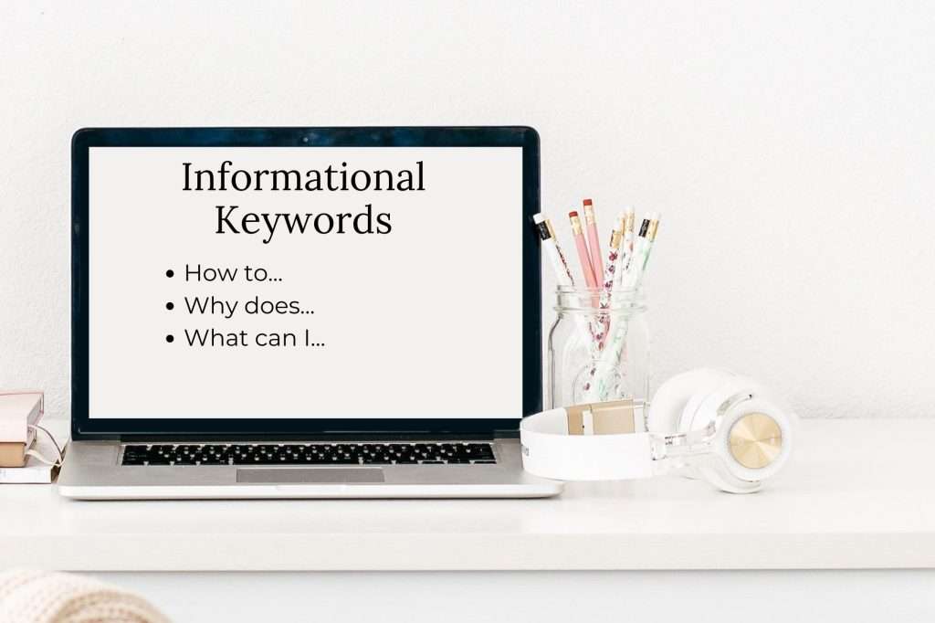 Open laptop with the words "Informational keywords: How to...
Why does...
What can I..." Written on the screen. Pencils in a clear jar to the right and a pair of white and rose gold headphones in front of the jar. 
