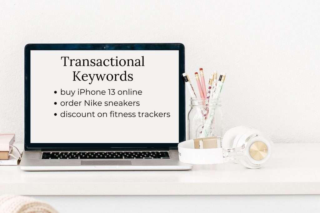 laptop on a white desk with white wall behind it. On the screen reads "Transactional Keywords: buy iPhone 13 online
order Nike sneakers
discount on fitness trackers" beside the laptop is a jar filled with pencils. In front of the jar is a white and rose gold headset.