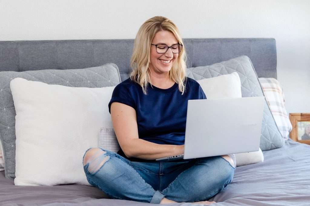 blonde woman with glasses wearing a blue top sitting on a bed with a laptop on her knees
