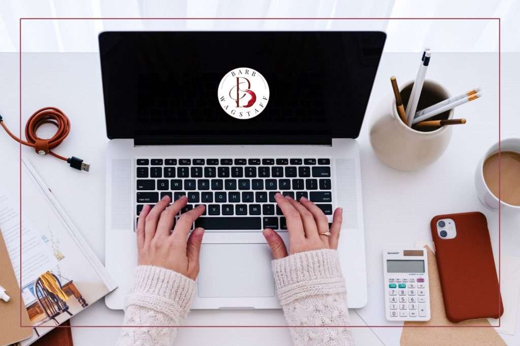 laptop with woman's hands on the keyboard. Calculator, pencils, and cellphone is on the desk. Barb Wagstaff logo is displayed on the laptop screen