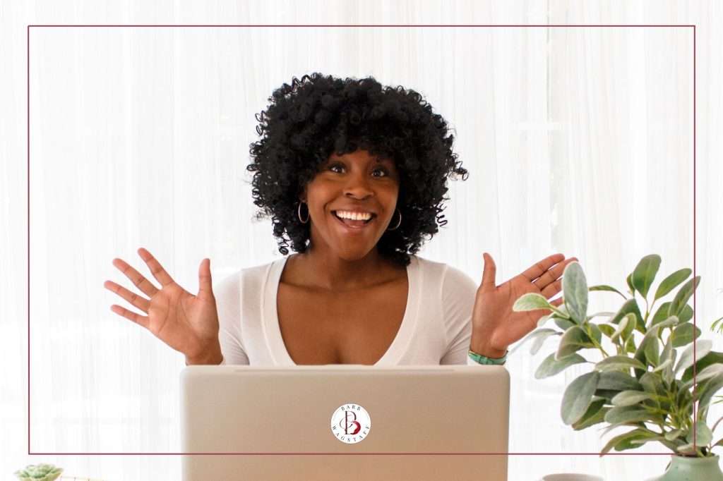 African woman in front of white curtain. Green plant on the right of the image. Laptop screen in front of her.