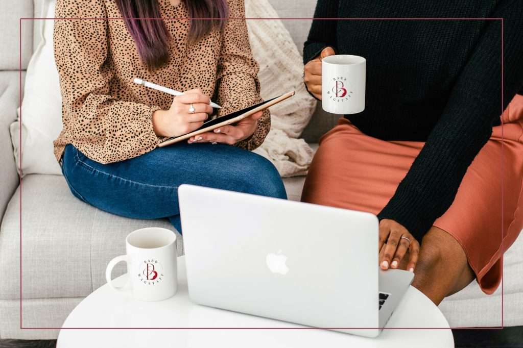 woman with a pen and ipad in her hand taking notes while other woman has coffee cup and is typing on her laptop